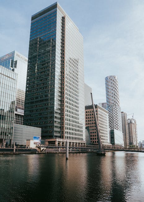 A tall, modern skyscraper with a predominantly glass exterior reflecting the sky and surrounding buildings, situated alongside a river or canal in an urban area. The building features a grid-like pattern of window panels with dark frames, and its facade shows a mix of dark tinted glass and light-colored concrete or metal accents. To the right, there are additional high-rise buildings, including a cylindrical tower with a distinctive patterned surface, and other structures with varying architectural styles, some under construction with cranes visible. The scene is illuminated by natural daylight, with a clear sky showing faint clouds. The water in the foreground is calm, creating a mirror-like reflection of the buildings, emphasizing the city's modern skyline. The area appears to be a commercial district, with clean surroundings and no visible rubbish or waste management equipment, exemplifying an environment where private or independent waste disposal might be employed by businesses or building management companies, such as Rubbish Removal Canary Wharf, to maintain cleanliness and manage waste efficiently.