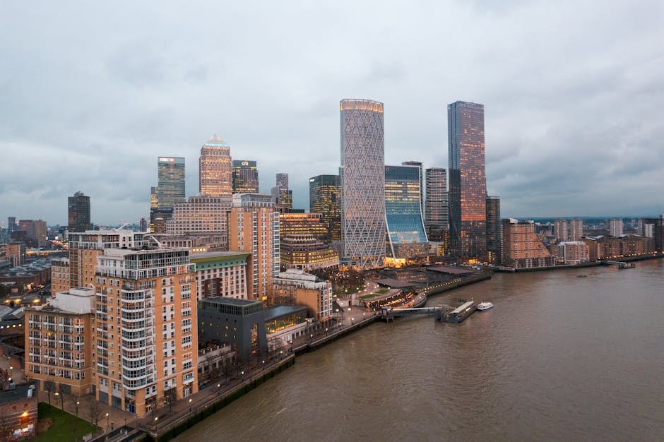 A panoramic view of the Canary Wharf financial district at dusk, featuring a cluster of modern high-rise office buildings with glass facades, some reflecting the orange and pink hues of the setting sun. The tallest skyscrapers include a distinctive cylindrical tower with a lattice-like exterior, and a rectangular tower with a dark reflective surface. In the foreground, a riverside walkway runs parallel to the Thames, with a few small boats moored along the quay. Surrounding the riverbank are mid-rise residential buildings constructed from brick and concrete, with large windows and balconies. The overcast sky casts a soft, diffuse light over the scene, creating a calm but busy urban atmosphere. This image illustrates the contrast between the historic and contemporary architecture of London’s riverside area, often involved in private waste management and alternative rubbish removal services, such as those provided by Rubbish Removal Canary Wharf.