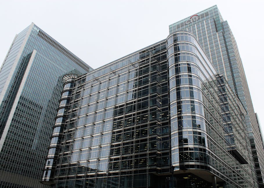 A modern, multi-story office building with a curved glass facade reflecting the surrounding cityscape, located in an urban area with tall skyscrapers. The building features tinted glass windows framed by dark metal, creating a sleek and contemporary appearance. The structure's exterior showcases horizontal and vertical lines formed by the window frames, with the rounded edges giving it a distinctive look. The scene is captured from a low angle, emphasizing the building's height against a gray, overcast sky. Adjacent high-rise buildings are visible on either side, with the one on the left displaying a lighter-colored glass exterior with vertical paneling, while the one behind has a similar glass and metal construction. The environment suggests a busy financial district, where such buildings are often associated with business offices and commercial activities, supporting services like private waste collection and rubbish removal for dense urban developments, such as those managed by Rubbish Removal Canary Wharf.