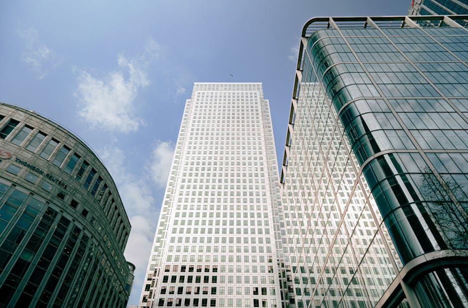 A low-angle view of a modern cityscape featuring three high-rise office buildings with reflective glass and steel facades. The central building has a white grid-patterned exterior with numerous small square windows, rising prominently into a partly cloudy blue sky. To the left, a rounded building with a curved glass frontage and vertical stone fins displays a name and logo, while the building on the right has a sleek, curved glass exterior with horizontal white metal trim. The scene is composed to highlight the architectural details and clean lines typical of contemporary commercial developments, with reflections of the neighboring structures visible on the glass surfaces. The image captures the urban environment associated with business districts that often require professional rubbish removal and private waste handling services, such as those offered by Rubbish Removal Canary Wharf, in a context aligned with office rubbish collection and on-site clearance activities.