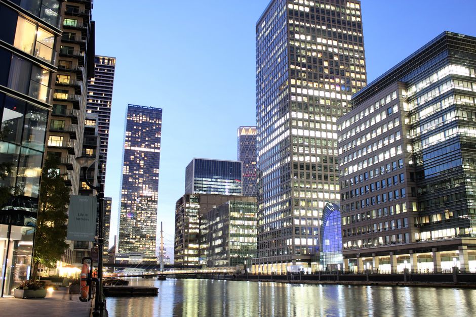 A modern, multi-story office building with a curved glass facade reflecting the surrounding cityscape, located in an urban area with tall skyscrapers. The building features tinted glass windows framed by dark metal, creating a sleek and contemporary appearance. The structure's exterior showcases horizontal and vertical lines formed by the window frames, with the rounded edges giving it a distinctive look. The scene is captured from a low angle, emphasizing the building's height against a gray, overcast sky. Adjacent high-rise buildings are visible on either side, with the one on the left displaying a lighter-colored glass exterior with vertical paneling, while the one behind has a similar glass and metal construction. The environment suggests a busy financial district, where such buildings are often associated with business offices and commercial activities, supporting services like private waste collection and rubbish removal for dense urban developments, such as those managed by Rubbish Removal Canary Wharf.