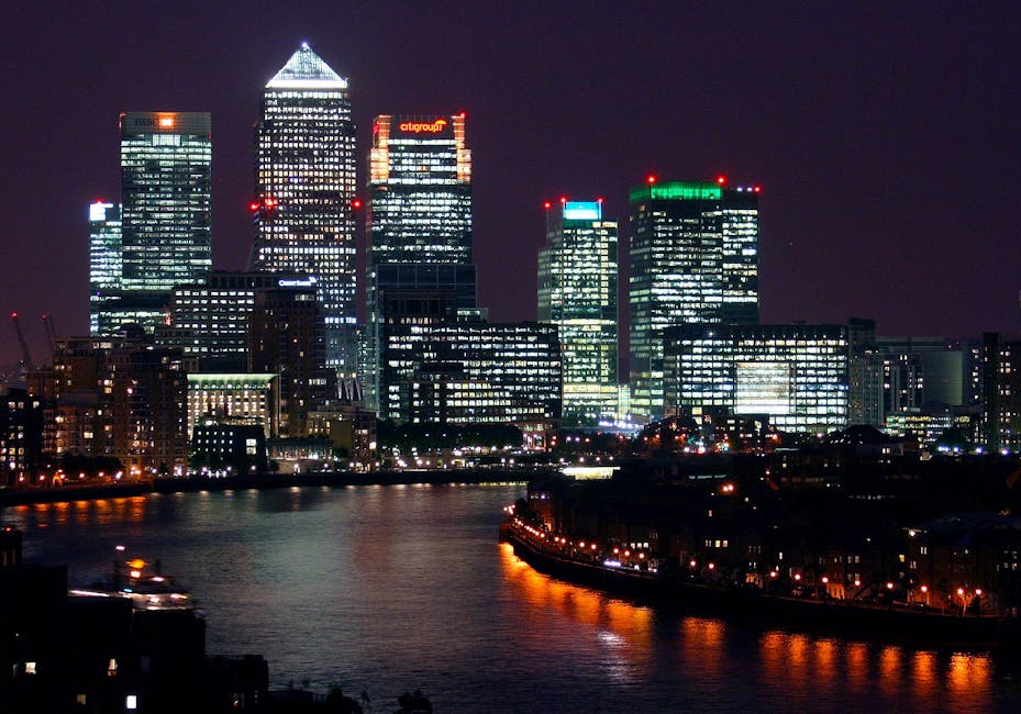 A cityscape featuring a tall modern skyscraper with a predominantly glass facade, reflecting the surrounding buildings and sky, situated along a river or waterway. The skyscraper has a sleek, rectangular profile with a darker, tinted upper section and a lighter, grid-patterned lower section. To the right, there are other high-rise buildings of various architectural styles, including one with a distinctive white, perforated cylindrical design and another with a curved, brown facade. The buildings are densely packed, forming a typical urban financial district scene. In the foreground, calm water reflects the buildings and sky, with a low wall or railing along the water's edge. The overall environment appears clean and modern, with natural daylight illuminating the scene and a slightly cloudy sky overhead. The setting suggests an area where potential private or independent waste management services, such as rubbish removal, might operate within the vibrant city environment, supported by nearby infrastructure.