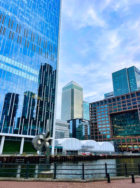 A cityscape view featuring a cluster of modern skyscrapers with reflective glass facades, including a tall blue-tinted building on the left, a cream-colored tower in the center, and a darker building with a grid-like window pattern on the right. In the foreground, there is a body of water with a black metal railing along the edge and a large, abstract metallic sculpture positioned on the water's edge. Behind the buildings, the sky is partly cloudy with a bright blue tone. The scene suggests an urban environment likely related to business and commercial activity, with the presence of static structures and water-based landscape. Although not visible in this specific image, such settings often involve various elements of rubbish removal and waste management, relating to sustainable urban development and independent waste disposal options, as seen in professional rubbish clearance services like Rubbish Removal Canary Wharf.