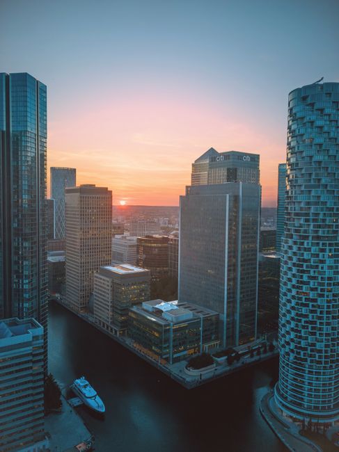 Aerial view of a modern cityscape at sunset showing several high-rise office towers with glass and concrete facades, positioned around a narrow canal or waterway. The buildings vary in height and architectural style, with some having reflective glass surfaces and others with textured stone or metal exteriors. One cylindrical tower with a patterned, brick-like finish stands prominently on the right, while rectangular structures dominate the scene. The waterway below is calm, with a small yacht positioned along the edge. The sky features a gradient of pastel colors, including pink, orange, and blue, with the sun partially visible on the horizon. The scene captures urban development in a dense financial district, suggestive of a location such as Canary Wharf, and emphasizes the modern architectural environment. This image could relate to private waste management or core infrastructure servicing large commercial buildings, as evidenced by the proximity of the structures and the urban environment, aligning with services offered by Rubbish Removal Canary Wharf for efficient, site-specific rubbish clearance.