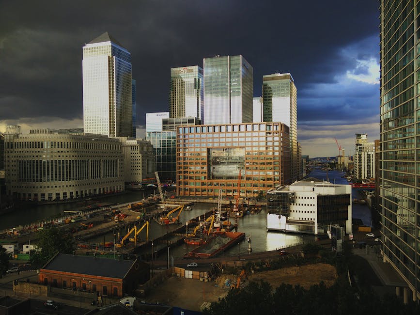 A cityscape view featuring a cluster of modern skyscrapers with reflective glass facades, including a tall blue-tinted building on the left, a cream-colored tower in the center, and a darker building with a grid-like window pattern on the right. In the foreground, there is a body of water with a black metal railing along the edge and a large, abstract metallic sculpture positioned on the water's edge. Behind the buildings, the sky is partly cloudy with a bright blue tone. The scene suggests an urban environment likely related to business and commercial activity, with the presence of static structures and water-based landscape. Although not visible in this specific image, such settings often involve various elements of rubbish removal and waste management, relating to sustainable urban development and independent waste disposal options, as seen in professional rubbish clearance services like Rubbish Removal Canary Wharf.
