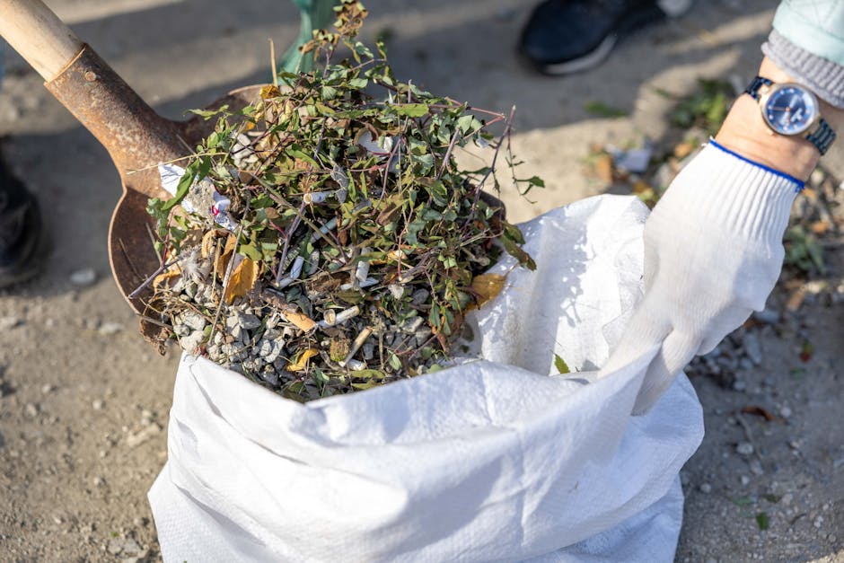 A close-up view of a gloved hand holding a large white sack filled with garden waste, including small twigs, dried leaves, soil, and some green plant stems. A rusty shovel is partly visible, positioned above the sack, with its blade covered in soil and organic debris. The background shows a gravel or dirt surface, suggesting an outdoor environment such as a driveway or garden area. The person wearing the glove has a wristwatch on, and another individual’s shoe is visible in the upper right corner, indicating active waste collection or clearance. The scene is illuminated by natural daylight, and the overall setting suggests a property cleanup, possibly handled through private waste removal services like Rubbish Removal Canary Wharf, providing an alternative to local authority rubbish collection.
